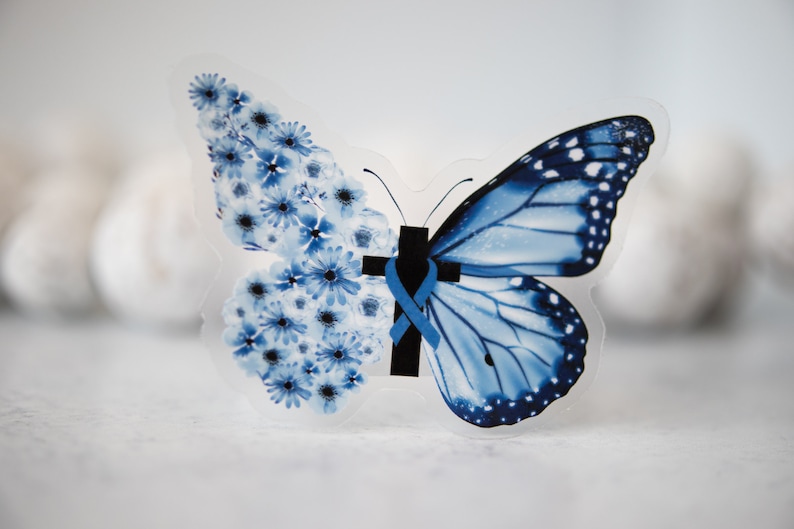 May include: A blue butterfly with a cross and a blue ribbon on its body. The butterfly is made of clear plastic and has a floral design on one wing.