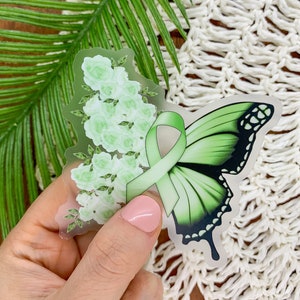 May include: A clear sticker with a green butterfly and a green ribbon. The butterfly has black markings and the ribbon is tied in a bow. The sticker is surrounded by white flowers.