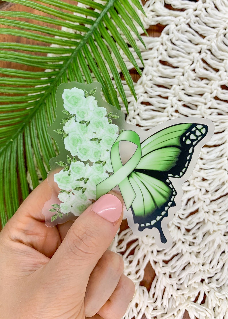 May include: A clear sticker with a green butterfly and a green ribbon. The butterfly has black markings and the ribbon is tied in a bow. The sticker is surrounded by white flowers.