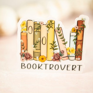 Pu&ograve; includere: Un adesivo con una pila di libri colorati con fiori e vegetazione. Il testo "BOOKTROVERT" &egrave; sotto i libri.