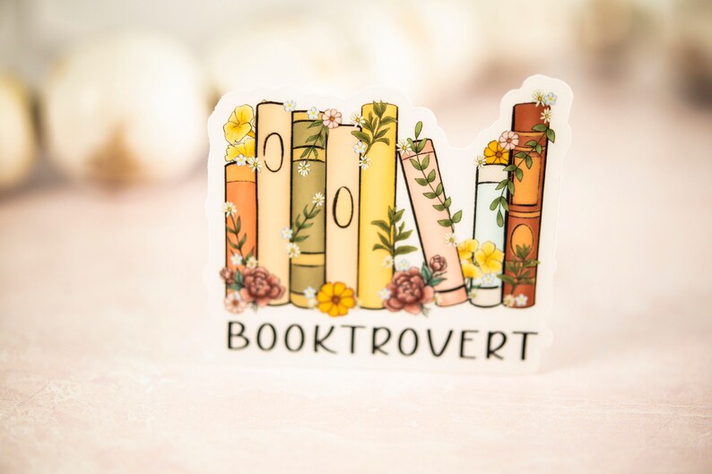 Pu&ograve; includere: Un adesivo con una pila di libri colorati con fiori e vegetazione. Il testo "BOOKTROVERT" &egrave; sotto i libri.
