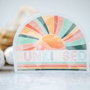 May include: A sticker featuring a retro sunburst design in shades of pink, orange, green, and yellow. The text "SUNKISSED" is printed in a bold, retro font.