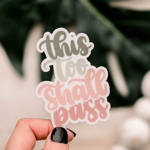 May include: A sticker with the text "This too shall pass" in a pink and gray gradient. The sticker is held in a hand with black nail polish.
