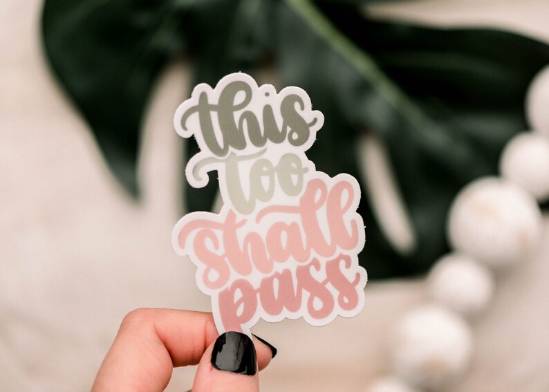 May include: A sticker with the text "This too shall pass" in a pink and gray gradient. The sticker is held in a hand with black nail polish.
