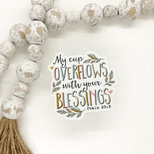 May include: A sticker with a white background and colorful text that reads "My cup overflows with your blessings" with a small heart on either side of the text. The text is in a handwritten font and is surrounded by small floral designs. The bottom of the sticker reads "Psalm 23:5".