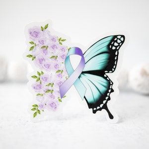 Pode incluir: Uma borboleta turquesa com marcas pretas e uma fita roxa com um centro azul claro. A borboleta é cercada por um grupo de flores roxas.