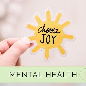 May include: A yellow sun sticker with the text "Choose Joy" written in black. The sticker is being held by a hand.
