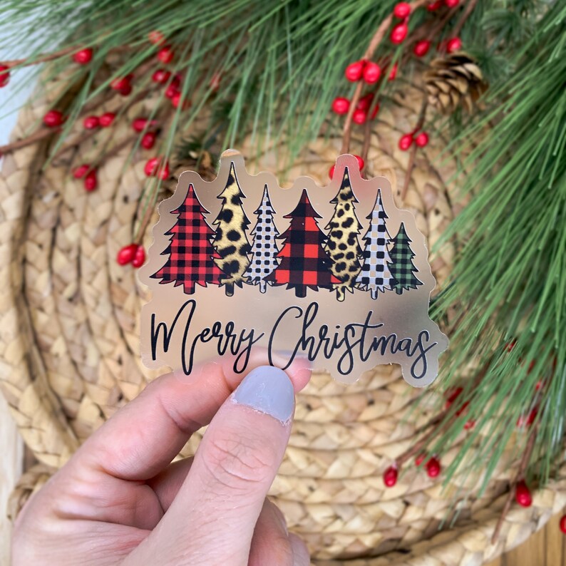 Puede incluir: Pegatina transparente con una fila de seis &aacute;rboles de Navidad en varios patrones, incluyendo cuadros, estampado de leopardo y lunares. Los &aacute;rboles son verdes, rojos, negros y blancos. El texto "Merry Christmas" est&aacute; escrito en cursiva negra debajo de los &aacute;rboles.