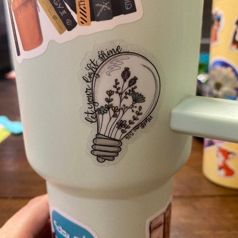May include: A light green tumbler with a sticker that says "Let your light shine" with a lightbulb design and flowers inside.