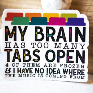May include: A white sticker with black text that reads "MY BRAIN HAS TOO MANY TABS OPEN 4 OF THEM ARE FROZEN & I HAVE NO IDEA WHERE THE MUSIC IS COMING FROM". The text is stylized to look like it was written with chalk. There are colorful tabs at the top of the sticker.