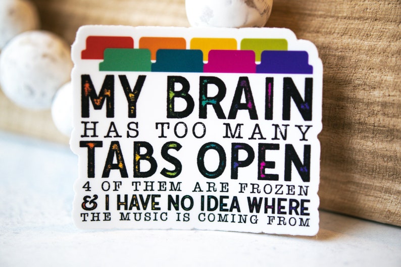 May include: A white sticker with black text that reads "MY BRAIN HAS TOO MANY TABS OPEN 4 OF THEM ARE FROZEN & I HAVE NO IDEA WHERE THE MUSIC IS COMING FROM". The text is stylized to look like it was written with chalk. There are colorful tabs at the top of the sticker.