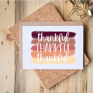 May include: A white card with the word "thankful" written in white cursive on a background of five horizontal stripes of color. The stripes are shades of red, orange, and yellow.