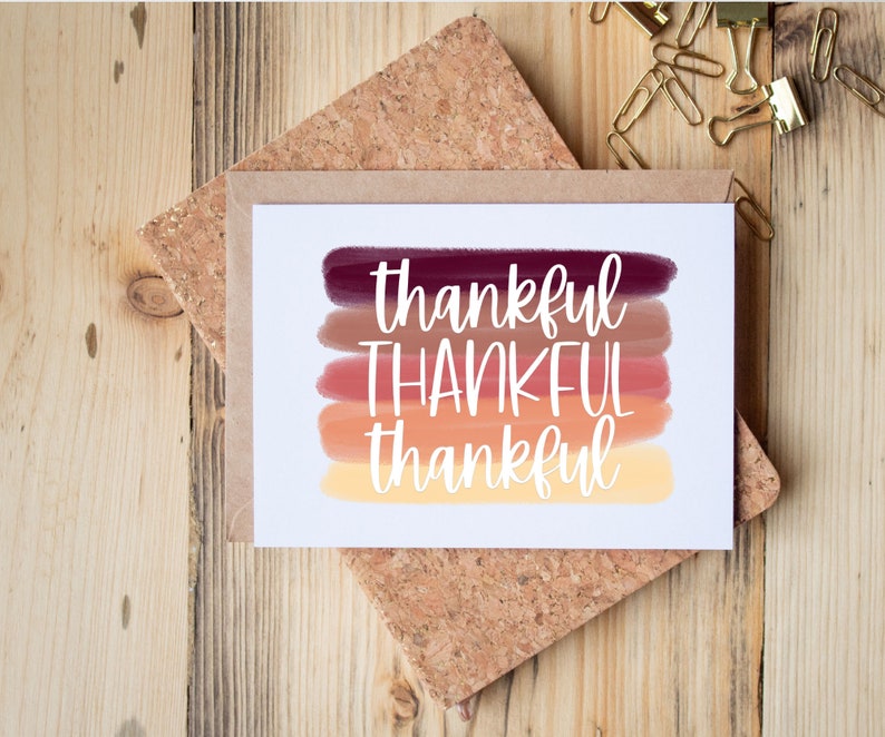May include: A white card with the word "thankful" written in white cursive on a background of five horizontal stripes of color. The stripes are shades of red, orange, and yellow.