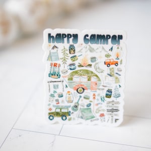 Pode incluir: Um autocolante com o texto "happy camper" em letras azuis e brancas. O autocolante apresenta uma ilustração de aquarela de uma autocaravana, montanhas, árvores e outros itens relacionados com o campismo.