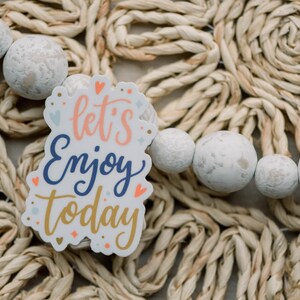 May include: White sticker with the text "let's enjoy today" in colorful lettering on a woven straw background with white beads.