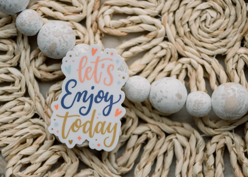 May include: White sticker with the text "let's enjoy today" in colorful lettering on a woven straw background with white beads.