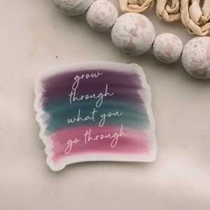 May include: A sticker with a watercolor background in shades of purple, teal, and pink. The sticker has the text "grow through what you go through" written in white cursive.