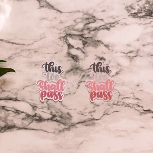 May include: Two pink and white stickers with the text "This too shall pass" on a marble background with pink flowers on either side.