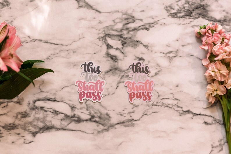 May include: Two pink and white stickers with the text "This too shall pass" on a marble background with pink flowers on either side.