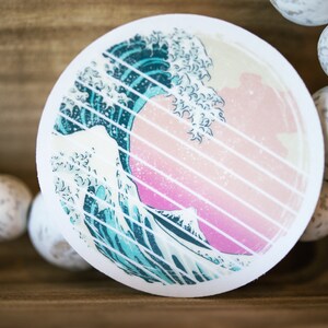 May include: A round sticker with a blue and white wave design. The wave is set against a pink and white striped background.