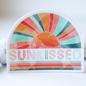 May include: A white sticker featuring a colourful retro sunburst design and the word "SUNKISSED" in a layered font. The sticker is on a white surface.