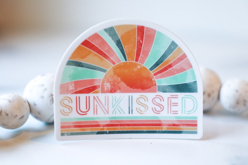 May include: A white sticker featuring a colourful retro sunburst design and the word "SUNKISSED" in a layered font. The sticker is on a white surface.