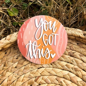May include: A round sticker with an abstract pink and orange design. The sticker says "You got this" in white cursive lettering with a small heart.