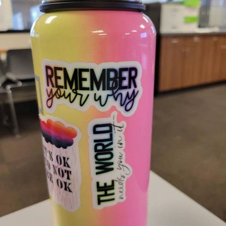 May include: A pink and yellow water bottle with multiple stickers. The stickers have inspirational quotes, including "Remember your why" and "The world needs you in it".