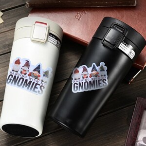 K&ouml;nnte beinhalten: Zwei Edelstahl-Reisebecher mit wei&szlig;er und schwarzer Oberfl&auml;che. Beide Becher haben einen Aufkleber mit einem Gnomendesign und dem Text "Hanging with my Gnomies".