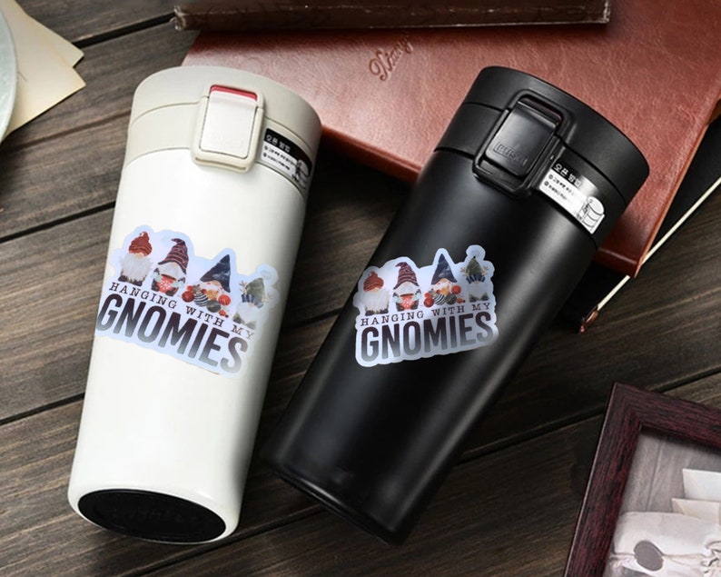 K&ouml;nnte beinhalten: Zwei Edelstahl-Reisebecher mit wei&szlig;er und schwarzer Oberfl&auml;che. Beide Becher haben einen Aufkleber mit einem Gnomendesign und dem Text "Hanging with my Gnomies".