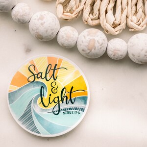 May include: A round sticker with a watercolor design of a sun rising over a wave. The sticker says "Salt & light" with the scripture reference "Matthew 5:13-16" underneath.