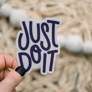 May include: A white sticker with the text "JUST DO IT" in purple lettering. The sticker is being held by a hand with black nail polish.