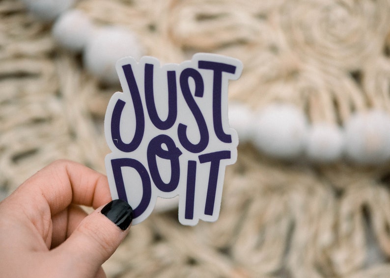 May include: A white sticker with the text "JUST DO IT" in purple lettering. The sticker is being held by a hand with black nail polish.