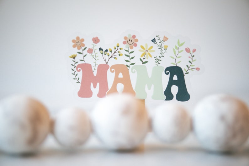 Pode incluir: Um sinal colorido de "MAMA" com flores e folhas. O sinal &eacute; feito de papel e tem um estilo retro e caprichoso.