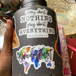 May include: A gray water bottle with a black lid, adorned with stickers. One sticker reads "Worry about NOTHING pray about EVERYTHING" and another features a colorful world map with the word "Wanderlust".
