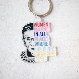 May include: A keychain featuring a black and white illustration of Ruth Bader Ginsburg with the text "Women belong in all places where decisions are being made" in rainbow colors.