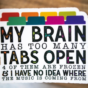 May include: A white sticker with black text that reads "MY BRAIN HAS TOO MANY TABS OPEN 4 OF THEM ARE FROZEN & I HAVE NO IDEA WHERE THE MUSIC IS COMING FROM". The sticker has a row of colorful tabs at the top.