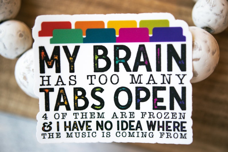 May include: A white sticker with black text that reads "MY BRAIN HAS TOO MANY TABS OPEN 4 OF THEM ARE FROZEN & I HAVE NO IDEA WHERE THE MUSIC IS COMING FROM". The sticker has a row of colorful tabs at the top.