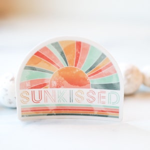 May include: A white sticker featuring a colourful rainbow sunburst design and the text "SUNKISSED" in a retro font.