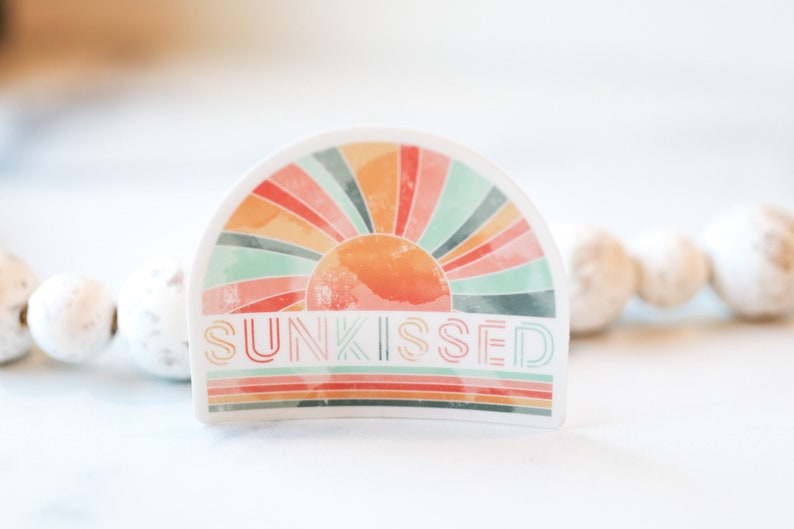 May include: A white sticker featuring a colourful rainbow sunburst design and the text "SUNKISSED" in a retro font.
