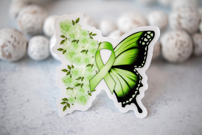 May include: Green butterfly sticker with a green ribbon and white flowers. The butterfly has black markings on its wings.