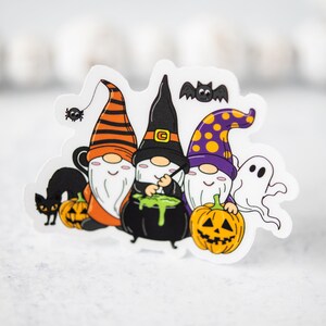 Puede incluir: Tres gnomos vestidos con trajes de Halloween, uno con un sombrero morado y amarillo, otro con un sombrero negro y otro con un sombrero a rayas naranja y negro. Los gnomos sostienen una caldera y calabazas. Hay un gato negro, una ara&ntilde;a y un fantasma en el fondo.