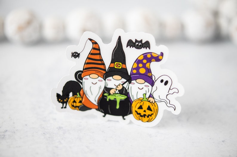Puede incluir: Tres gnomos vestidos con trajes de Halloween, uno con un sombrero morado y amarillo, otro con un sombrero negro y otro con un sombrero a rayas naranja y negro. Los gnomos sostienen una caldera y calabazas. Hay un gato negro, una ara&ntilde;a y un fantasma en el fondo.