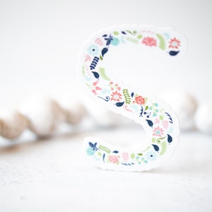 Könnte beinhalten: Ein weißer Papierschnitt des Buchstabens "S" mit einem floralen Muster in den Farben Rosa, Blau, Grün und Marine.