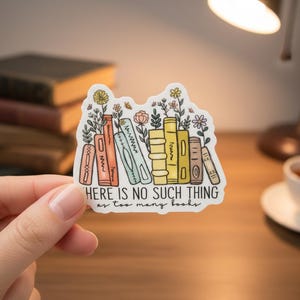 May include: A sticker featuring a stack of illustrated books with flowers and the text "HERE IS NO SUCH THING as too many books." The books are in various colors, and the sticker is held by a hand.