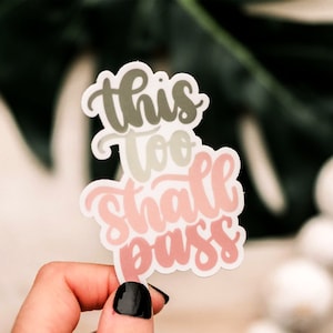 May include: A sticker with the text "This too shall pass" in a pink, green, and white color scheme. The sticker is held in a hand with black nail polish.