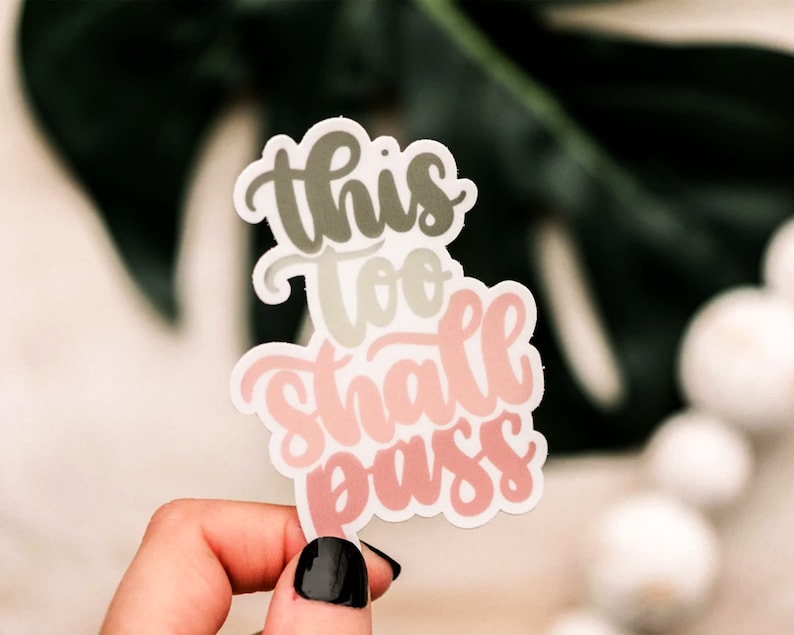 May include: A sticker with the text "This too shall pass" in a pink, green, and white color scheme. The sticker is held in a hand with black nail polish.