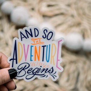 May include: A white sticker with the text "And so the adventure begins" in colorful lettering. The sticker is held in a hand.