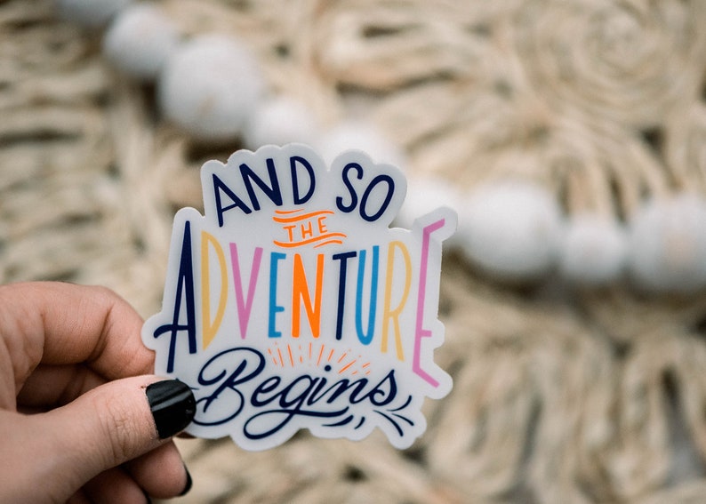 May include: A white sticker with the text "And so the adventure begins" in colorful lettering. The sticker is held in a hand.