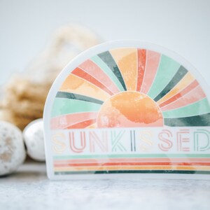 May include: A white sticker with a colourful sunburst design and the text "SUNKISSED" in a retro font. The sticker is half-circle shaped and has a watercolour effect.